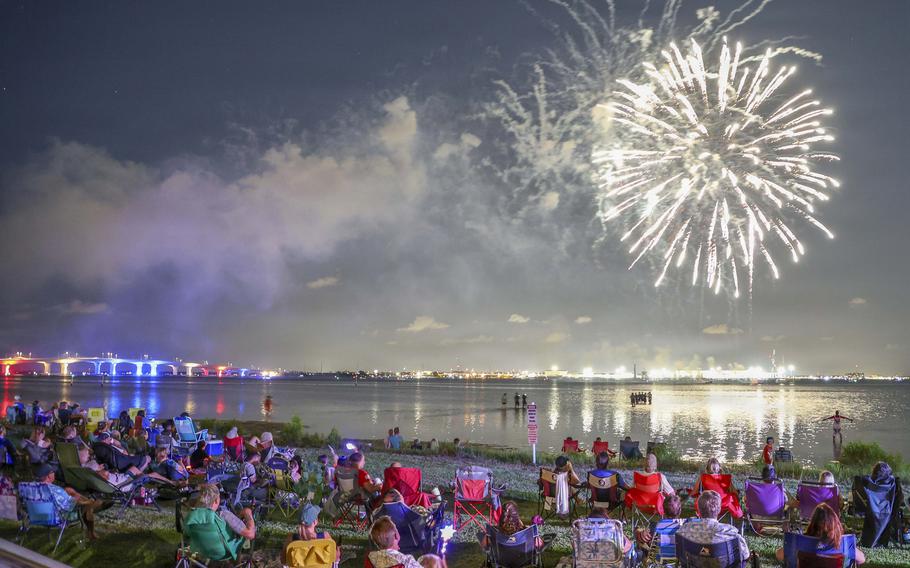 Crowds of people seated in lawn chairs and picnic blankets on the grass, watching fireworks explode over the water.