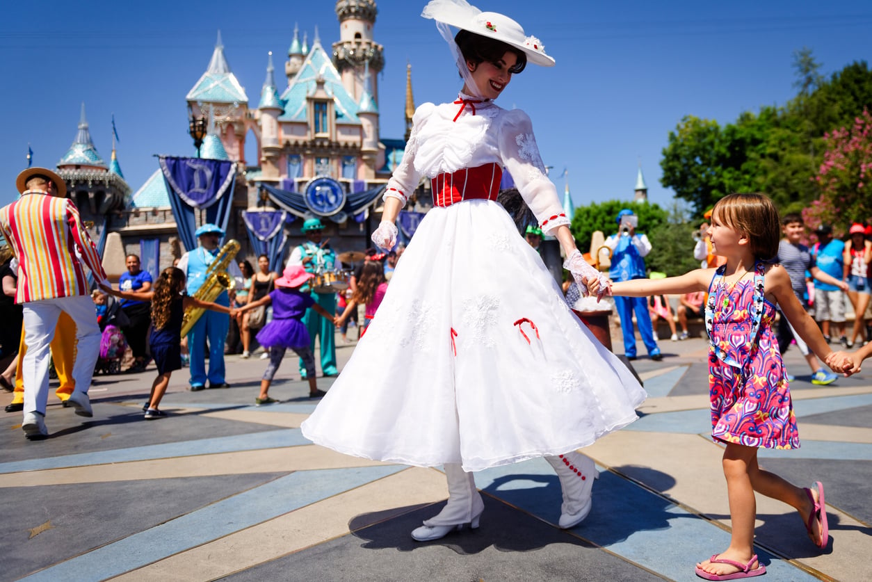 A woman in a white dress, resembling Mary Poppins, holds hands and dances with a smiling young girl at a colorful parade in front of a castle at one of the world’s most enchanting tourist attractions.