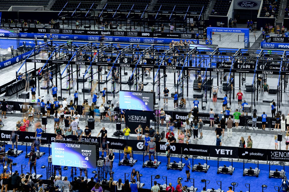 Crowd participating in a CrossFit event with multiple workout stations and branding displays in a large arena.