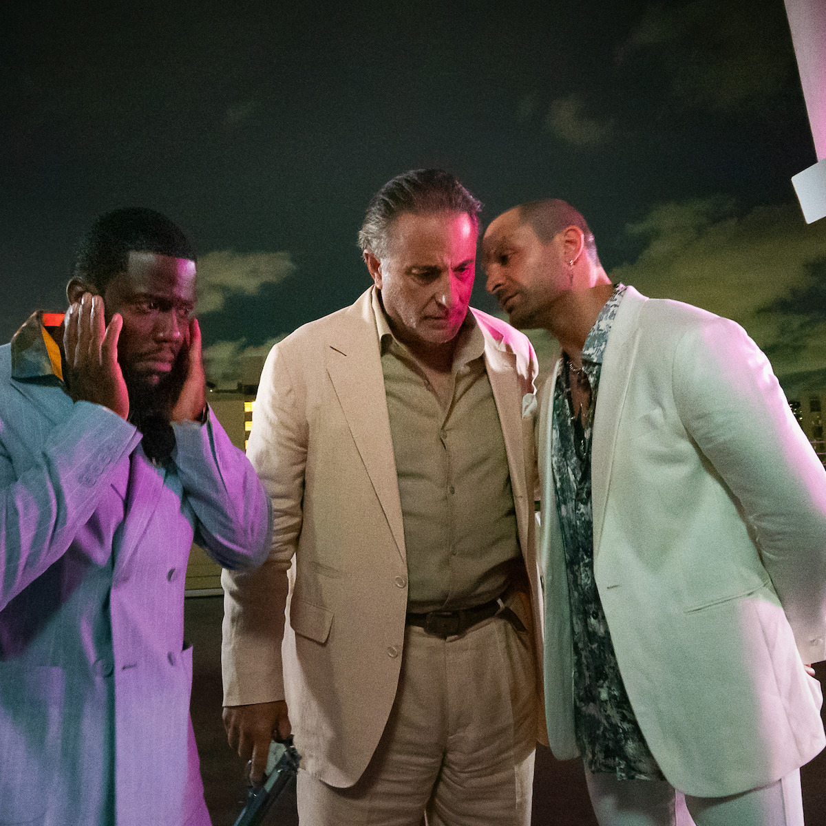 Three men in suits stand closely together outside at night, two in light-colored jackets, one covering his ears and the others in a tense conversation, under cloudy skies with city lights in the background.