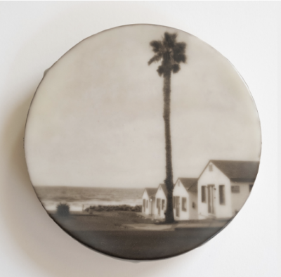 A circular sepia-toned image of a palm tree, small white building, and shoreline with encaustic media, © Ann Mitchell.