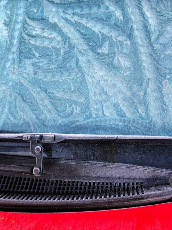 A car windshield covered in intricate frost patterns, resembling fern leaves, with a windshield wiper visible at the bottom and a glimpse of the red car hood.