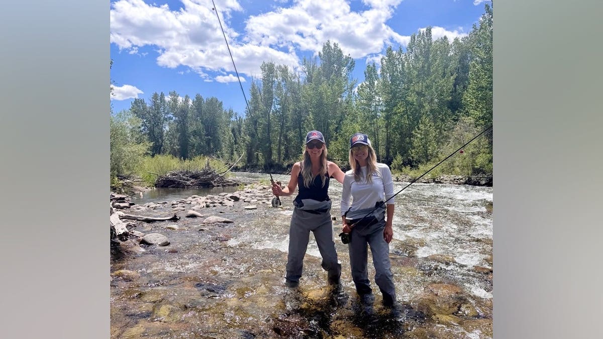Ali Larter stands in a mountain river wearing chest waders and holding a fly rod alongside a friend during a Fourth of July getaway.