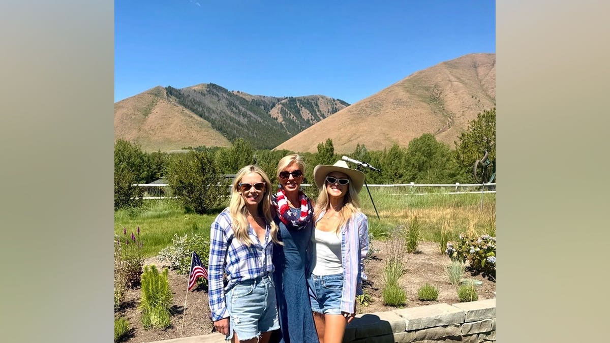 Ali Larter poses with two friends in front of a mountain landscape while wearing denim shorts, a white tank top, a striped button-down shirt and a white cowboy hat.