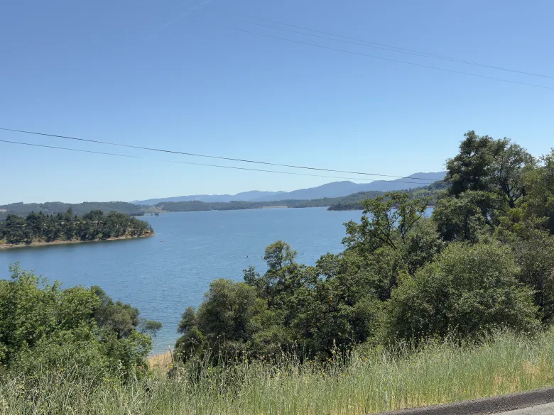 A serene lake surrounded by lush green trees and distant hills under a clear blue sky.