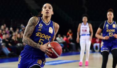 A female basketball player in a blue uniform with the number 5 holds the ball, preparing to shoot, while two other players stand behind her on the court.