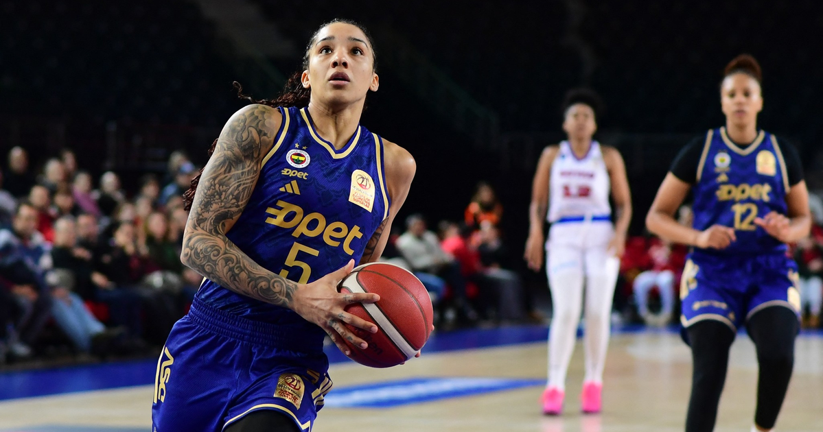 A female basketball player in a blue uniform with the number 5 holds the ball, preparing to shoot, while two other players stand behind her on the court.