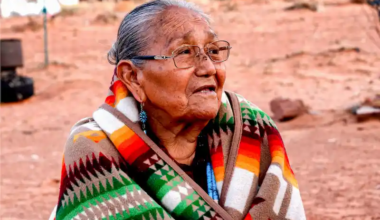 A Native American older woman, wearing glasses, turquoise earring, and a colorful tribal blanket, sits outside in the desert.