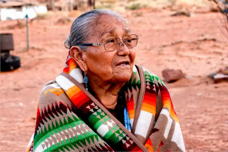 A Native American older woman, wearing glasses, turquoise earring, and a colorful tribal blanket, sits outside in the desert.