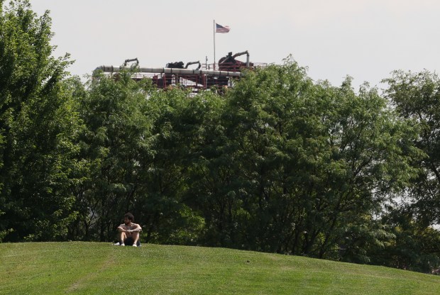 Pipes that connect silos at MAT Asphalt are visible from across the 2000 block of West Pershing Road at the south end of McKinley Park as area resident Cuitlahuac Arriaga rests on a hill, July 23, 2026, in Chicago. (John J. Kim/Chicago Tribune)