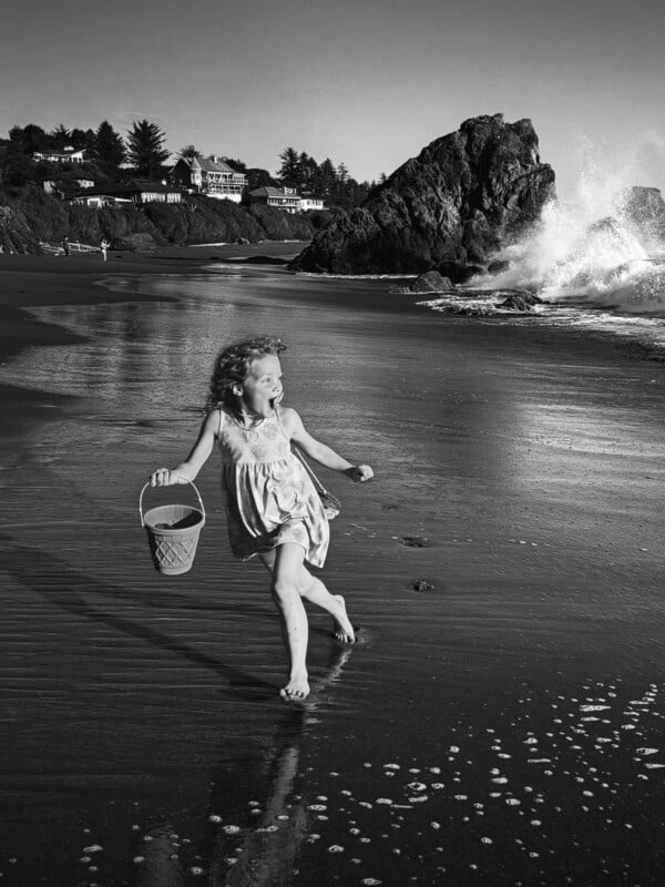 A young girl with curly hair runs barefoot on a beach, holding a bucket. Waves crash against rocks in the background, and houses sit on a hill beneath trees. The scene is in black and white.