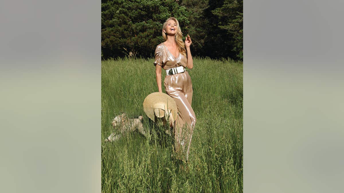 Christie Brinkley in a field smiling and holding a straw hat.