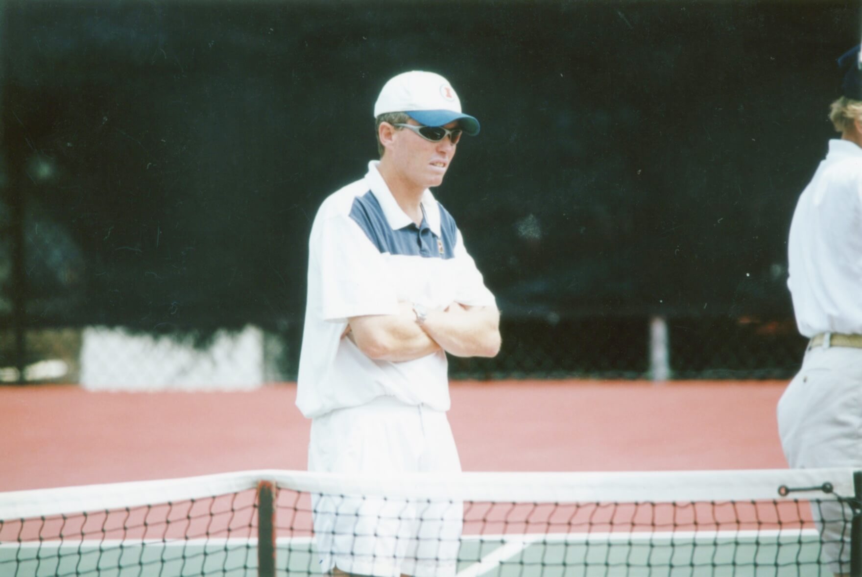 Craig Tiley stands on a green and red tennis court in 2003 wearing a cap and sunglasses. His arms are crossed.