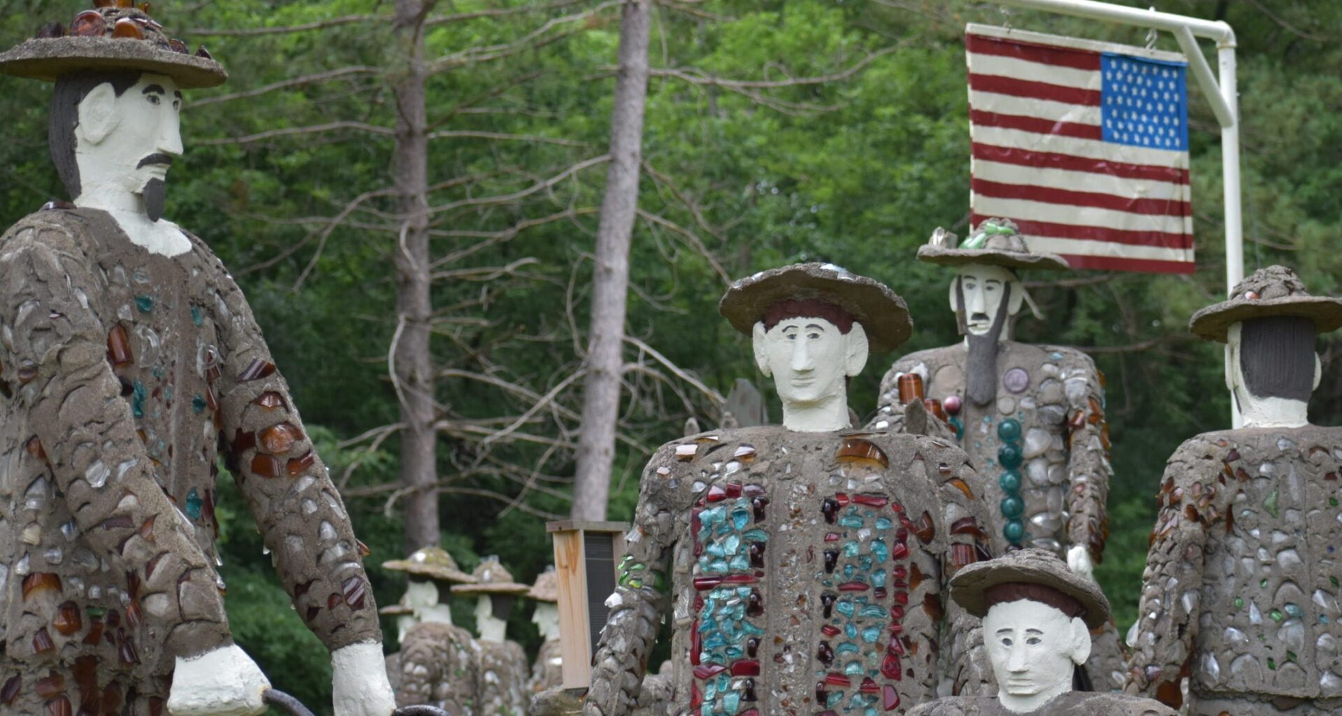 Several stone and glass mosaic statues of people stand outdoors near trees with an American flag displayed on a pole in the background.