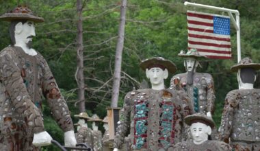 Several stone and glass mosaic statues of people stand outdoors near trees with an American flag displayed on a pole in the background.