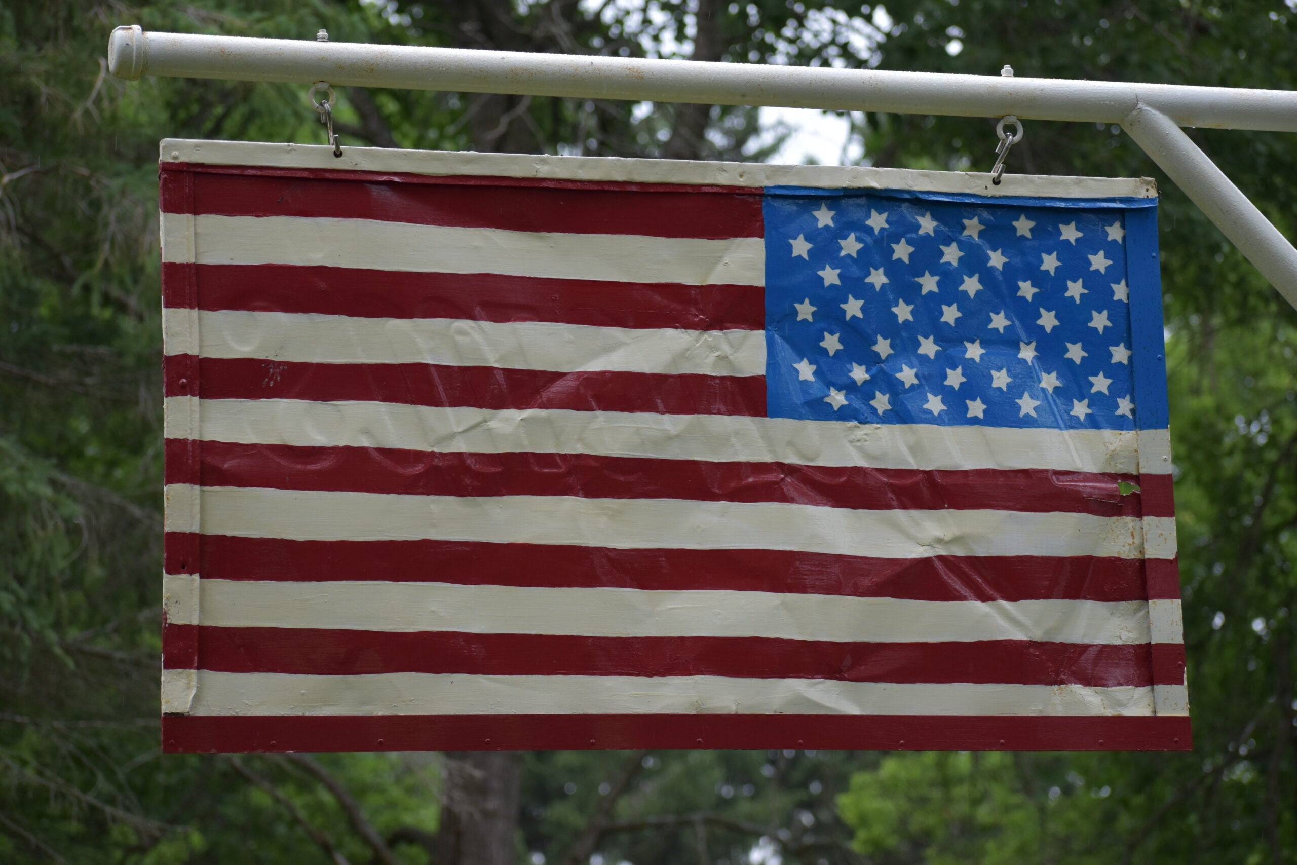 A U.S. flag is displayed upside down, with the stars in the lower right corner, hanging outdoors against a background of green trees.