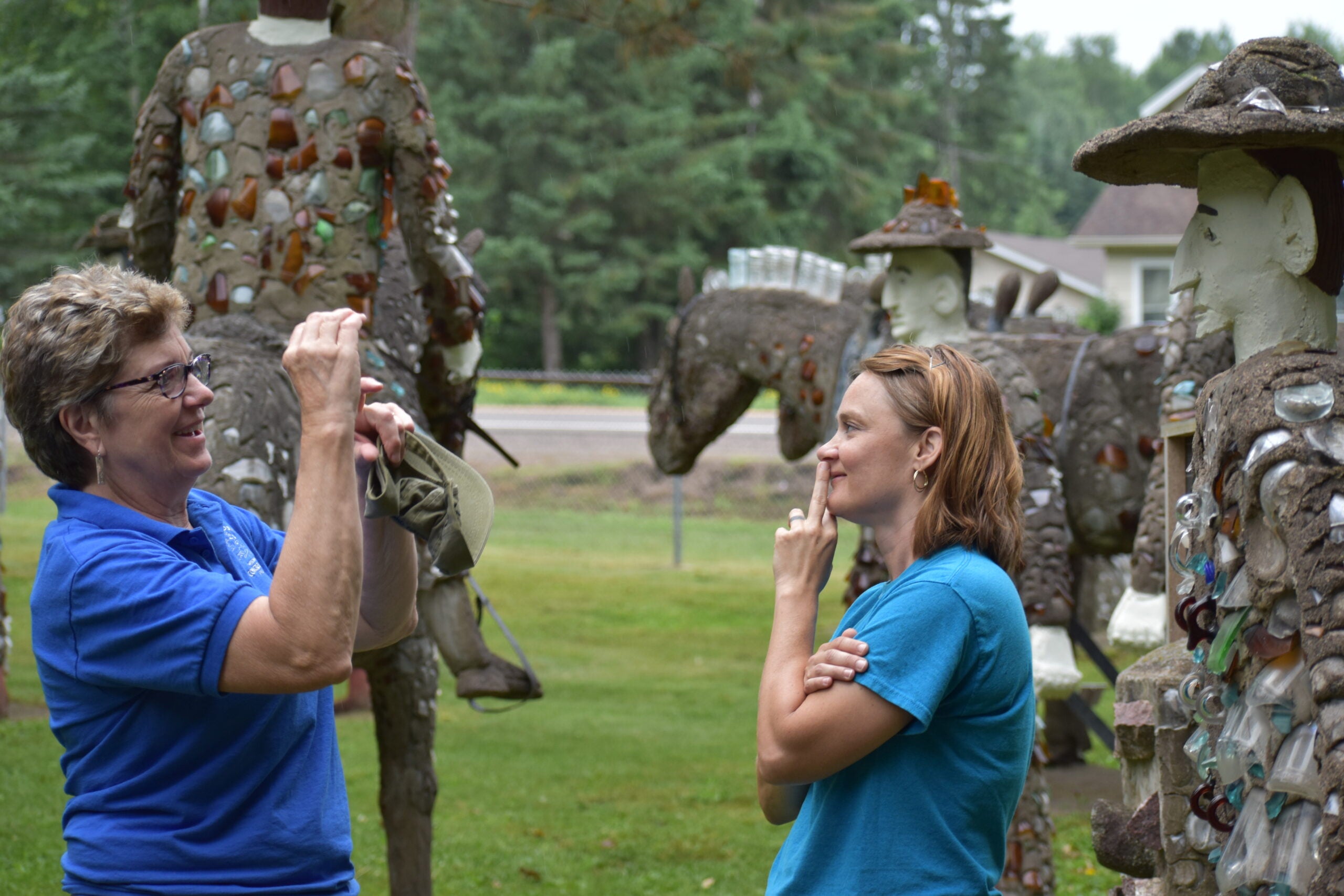 Two women stand outdoors, one taking a photo while the other poses with a finger to her nose, surrounded by statues decorated with glass fragments.