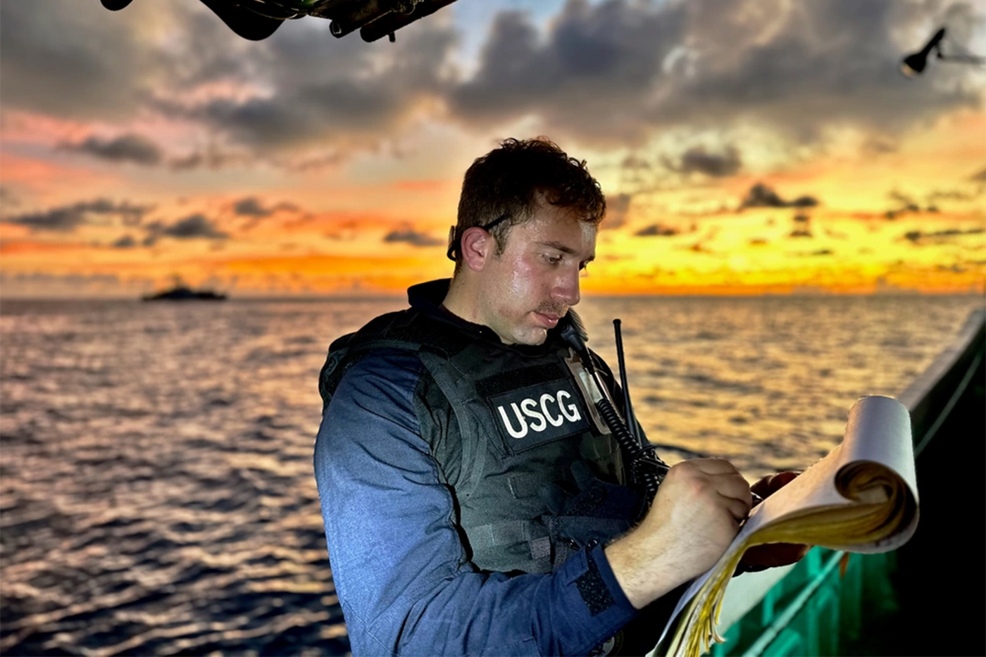 Drew LoVullo, in a U.S. Coast vest, writes on a paper while aboard a ship with the sunset in the background.