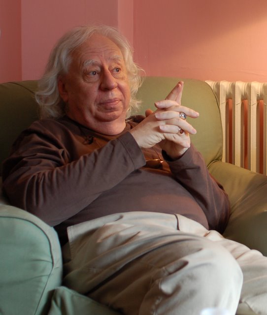 Edward Lucie-Smith sits in a green armchair in a cosy looking room