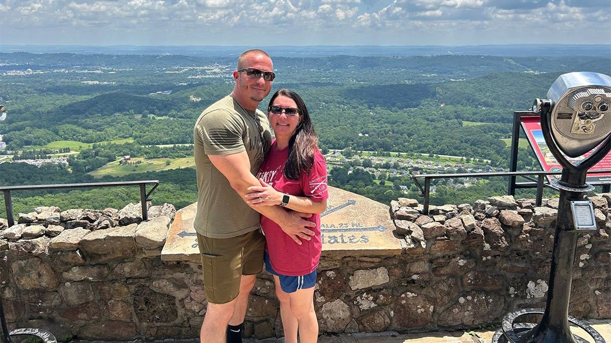 Justin Elrod, wearing sunglasses and an olive green T-shirt, and his wife Lena Elrod, wearing sunglasses and a red T-shirt, embrace while posing in front of a mountaintop view with a rock wall and coin-operated binoculars nearby.