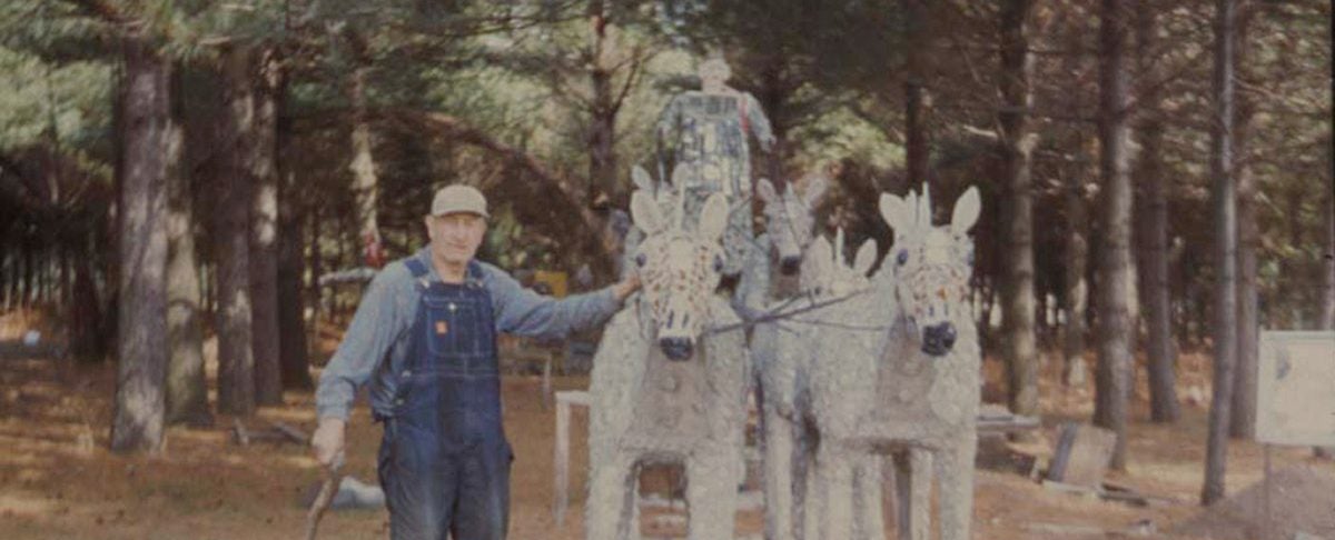 A man in overalls and a cap stands beside three life-sized, mosaic-covered horse statues in a wooded outdoor area.