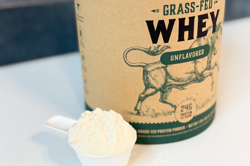 A canister of Heart & Soil Grass-Fed Whey Protein. A plastic scoop packed with the off-white powder sits next to it on the counter.