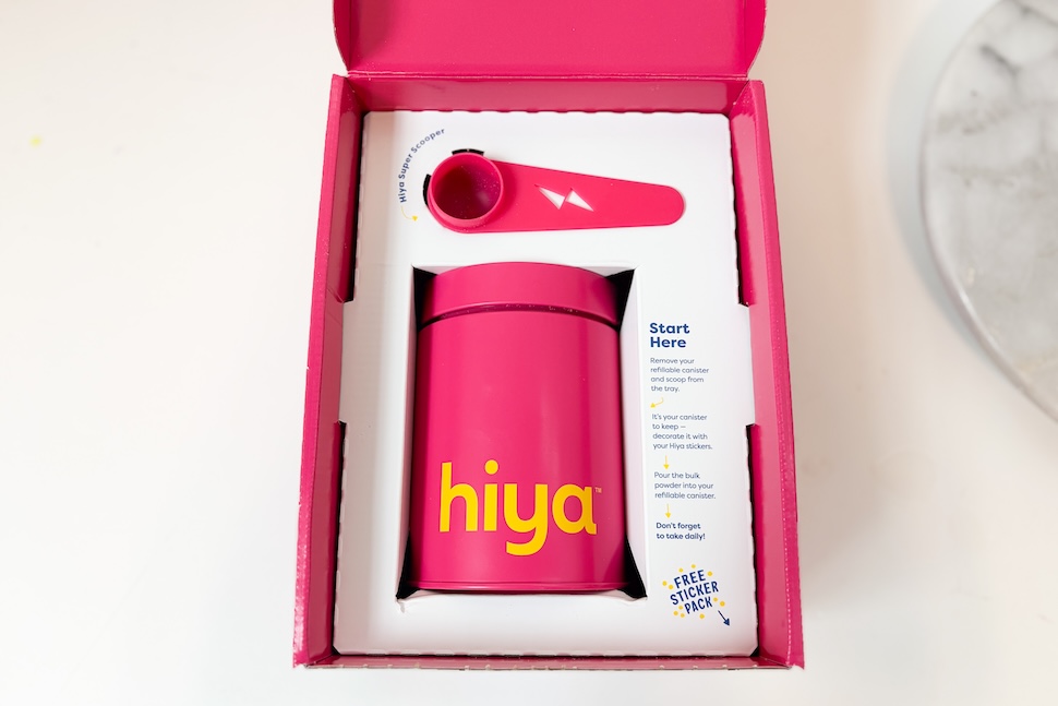 An open box of A box of Hiya Kids Daily Growth + Protein. It shows the canister of protein and scooper, both magenta in color.