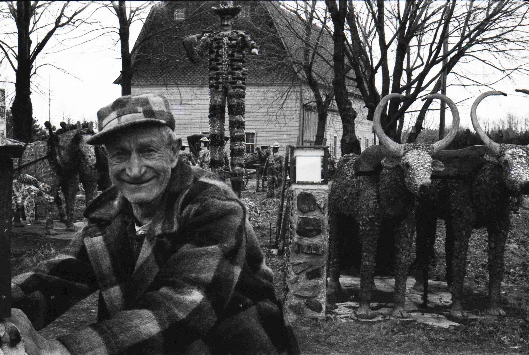 An older man in a plaid coat and cap smiles outdoors, standing near sculpted animal figures and tall statues in a yard with leafless trees and a house in the background.