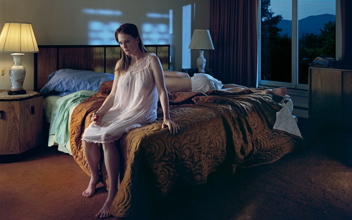A woman in a nightgown sits on the edge of a bed, looking down, while another person lies face down on the bed in a dimly lit bedroom.