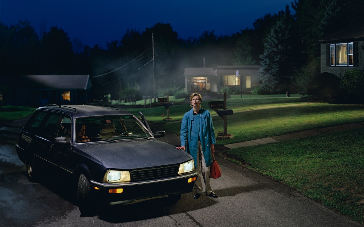 A person in a blue coat stands by an old station wagon at night in a suburban neighborhood, holding a red bag. The house windows are lit and another person sits inside the car.