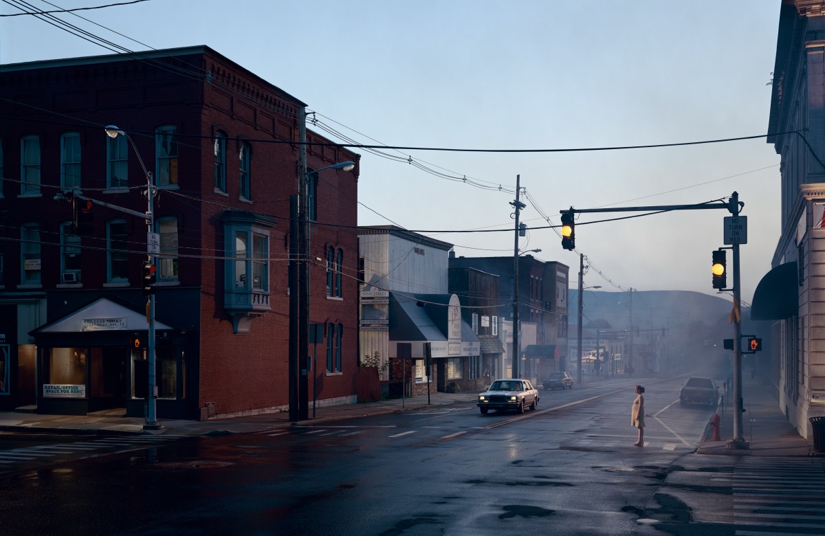A quiet, wet street in a small town at dawn, with a single car driving through an intersection as a person waits on the sidewalk.