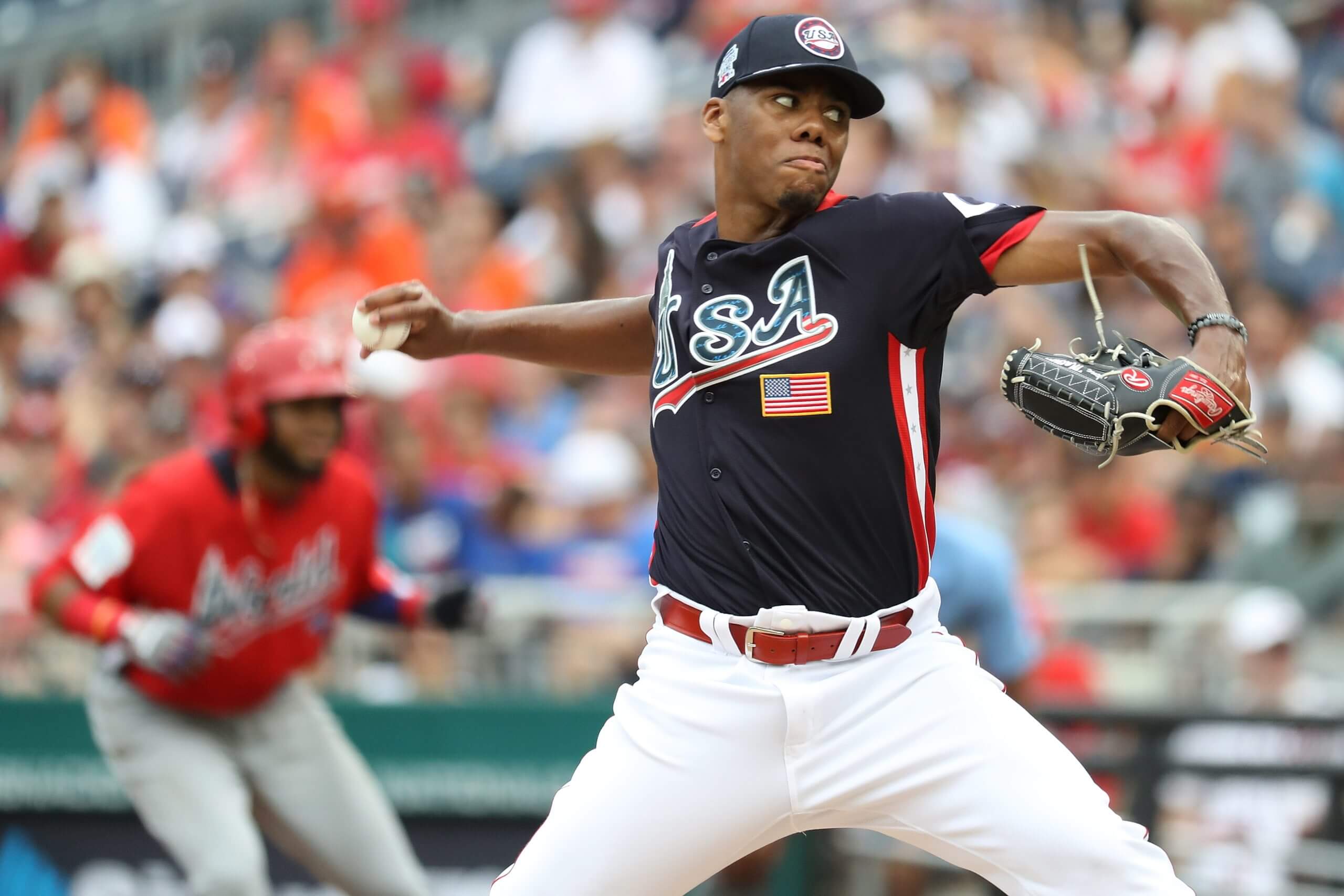 Hunter Greene delivers a pitch during the 2018 All-Star Futures Game with a base runner in a red helmet and a red jersey top in the background.