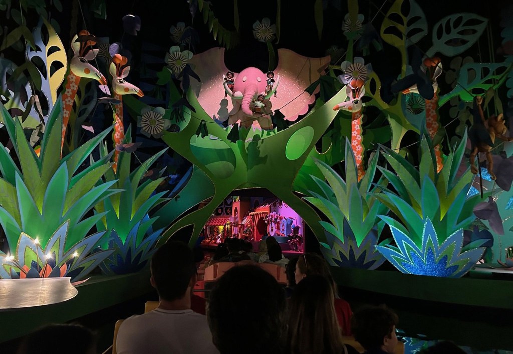 View from a boat ride of animatronic giraffes and an elephant with dolls in a jungle setting.