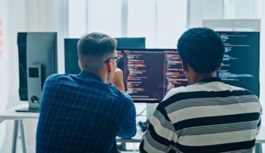 Two male software developers meeting on multiple computer screens displaying programming code, collaborating on project in office.