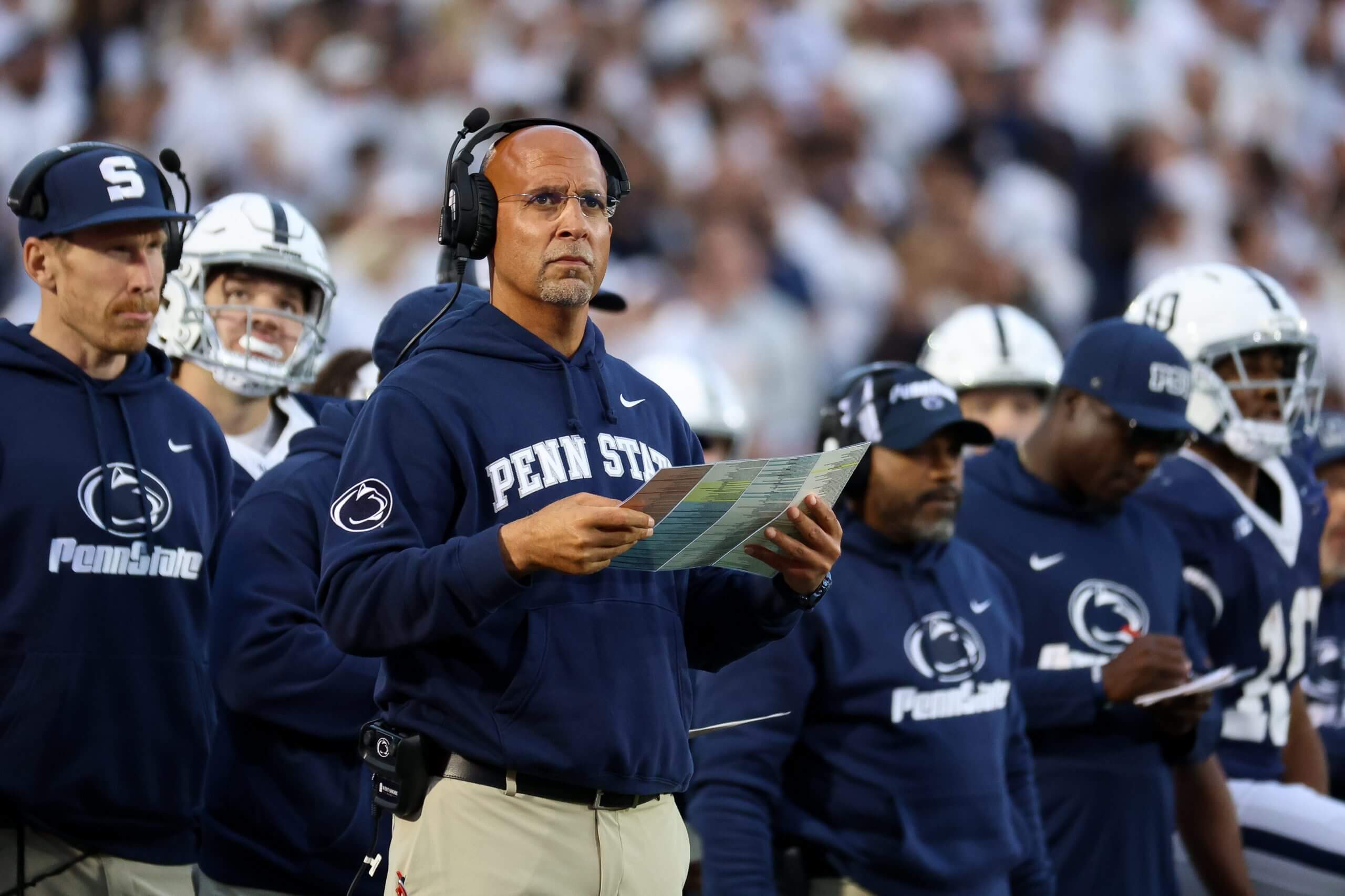 James Franklin holds a play sheet and looks toward the field with Penn State coaches and players behind him