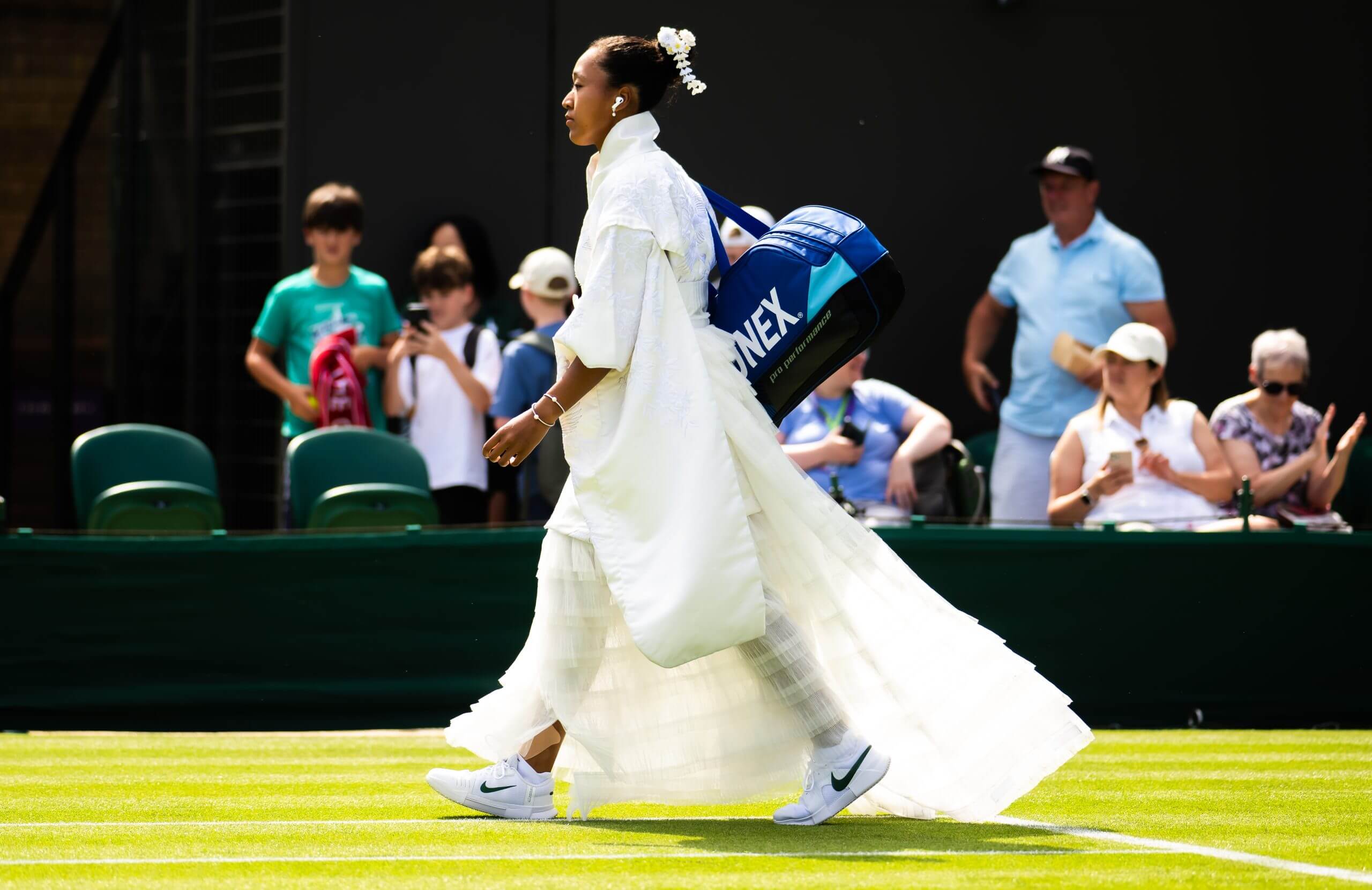 Naomi Osaka walks on a grass tennis court wearing a white ceremonial dress, plunging skirt and hairpiece.