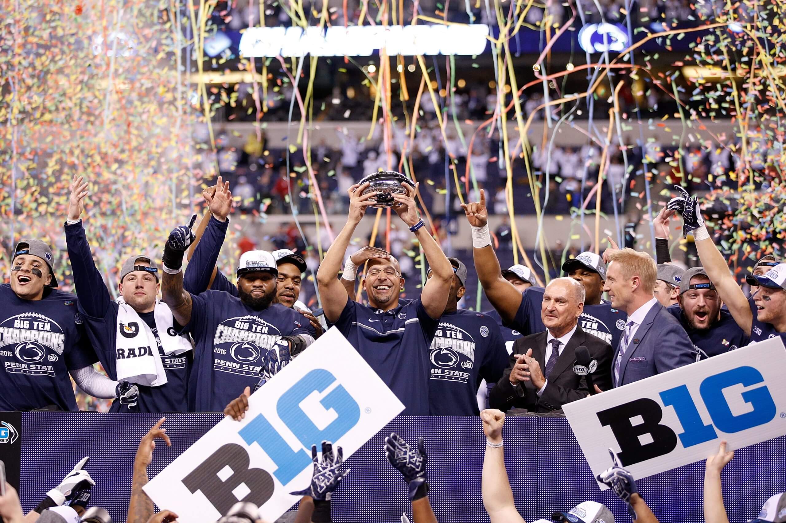 James Franklin holds up the Big Ten championship football trophy while confetti falls and Penn State players celebrate around him