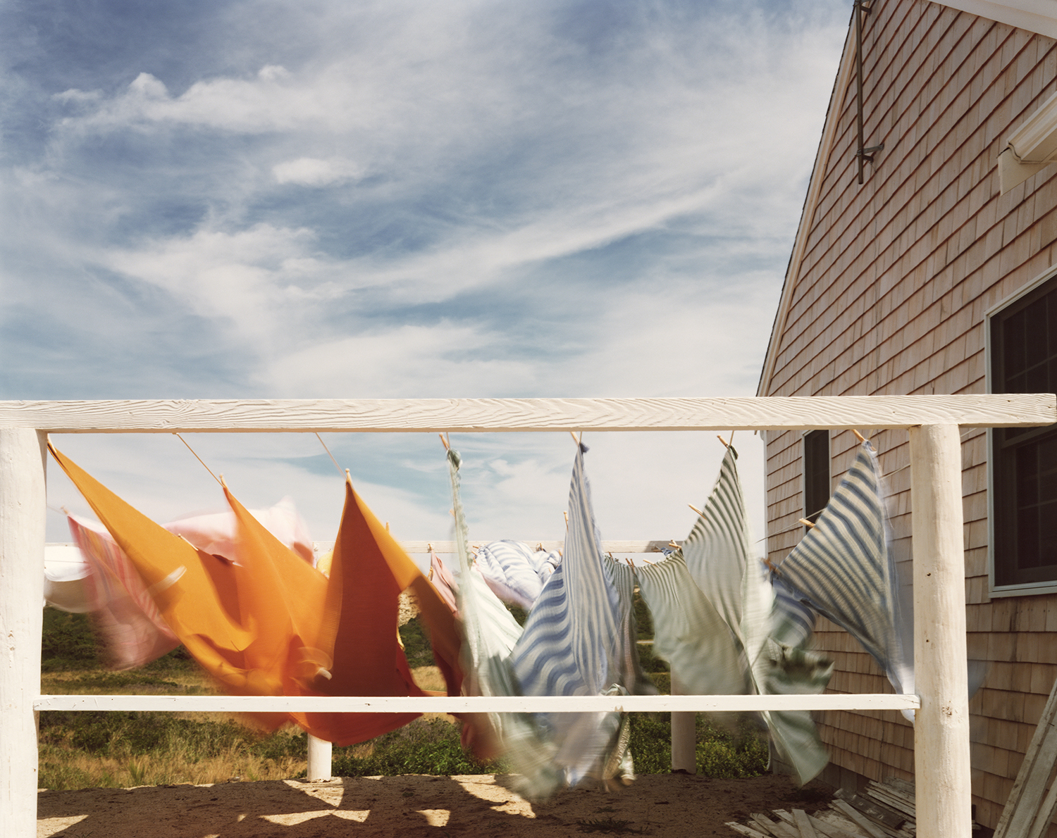 Brightly coloured washing, orange, pink and striped, billows on a line strung between white posts beside a shingled house under a wind-swept sky.