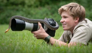 A smiling person lies on grass, holding a camera with a large lens while a snake is slithering over the lens; trees blur in the background.
