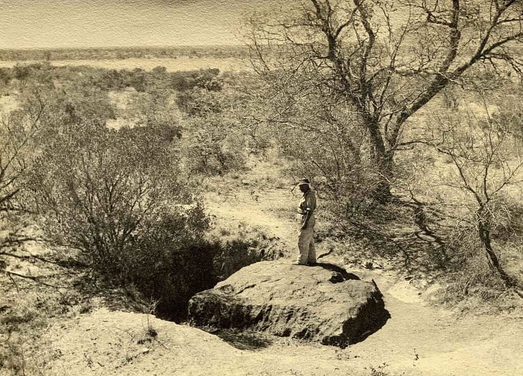 Sepia tone photo of a man standing above the Hoba meteorite partially excavated in a savanna landscape.