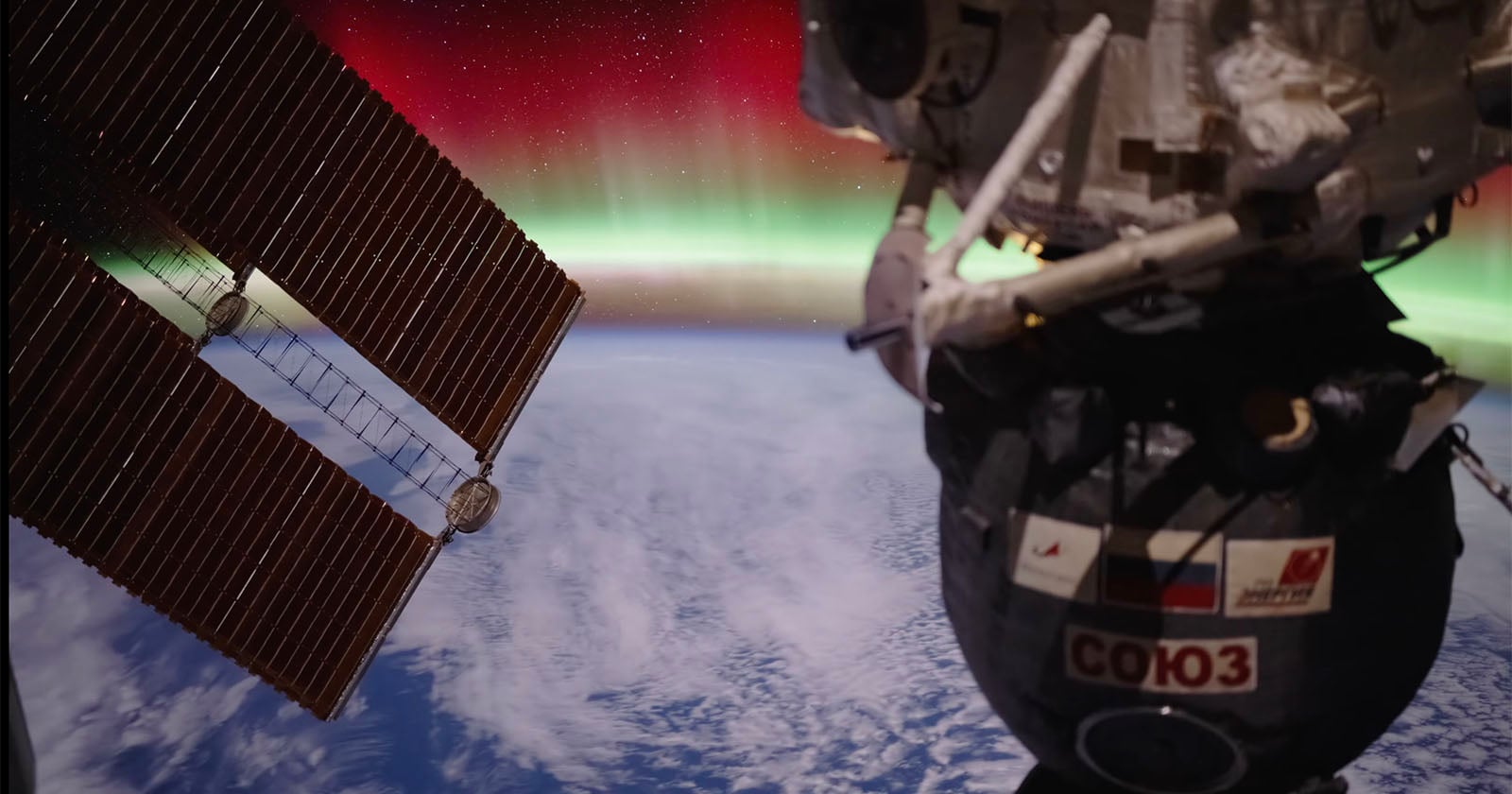 A view from the International Space Station shows a Russian Soyuz spacecraft and solar panels above Earth, with a glowing green and red aurora visible over the planet’s horizon.