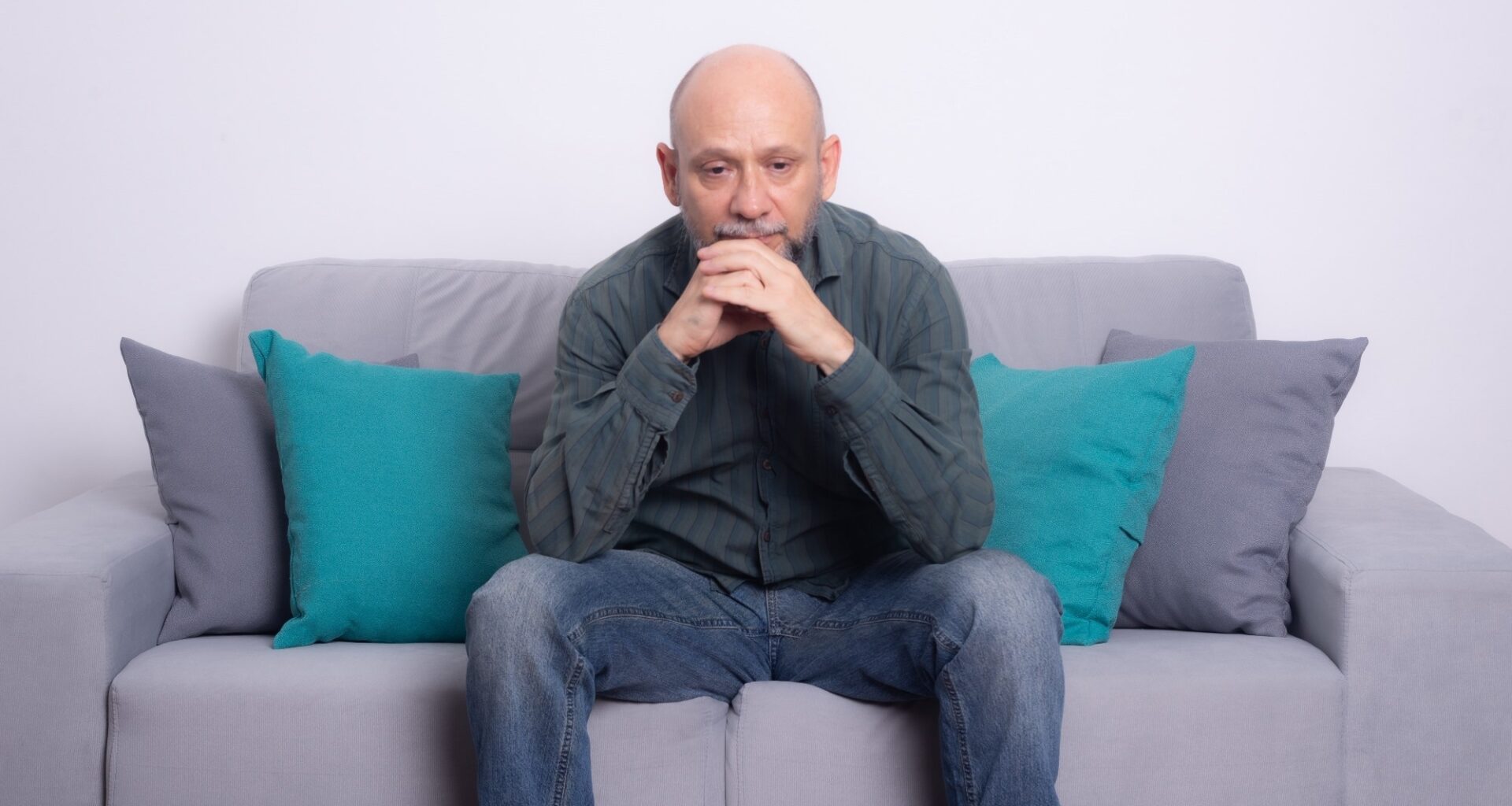 Bearded bald man wearing a long-sleeved shirt and jeans sitting on a sofa. Seriously looking down. Against a gray background.