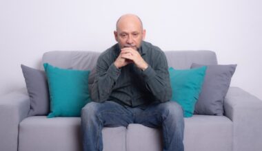 Bearded bald man wearing a long-sleeved shirt and jeans sitting on a sofa. Seriously looking down. Against a gray background.
