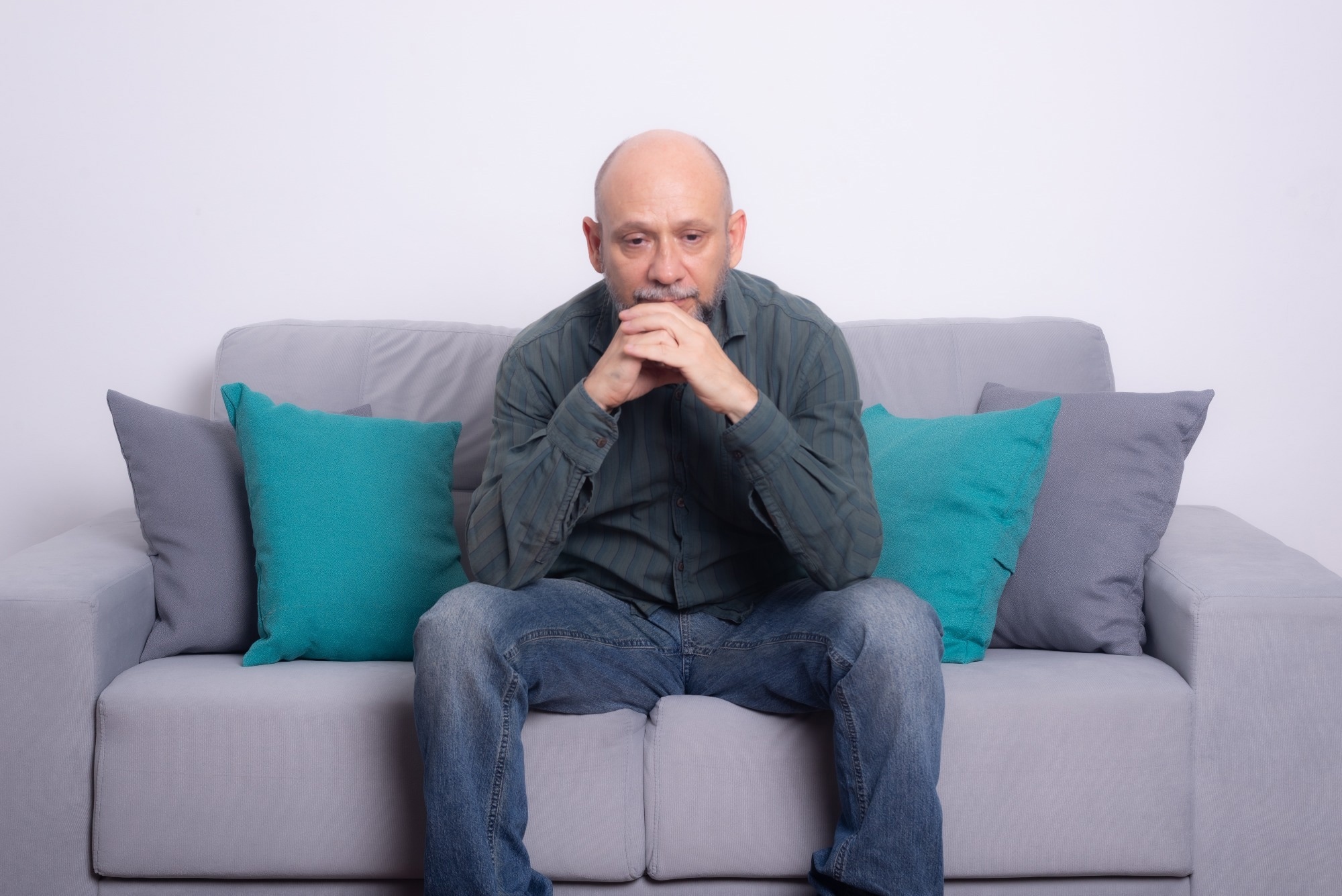 Bearded bald man wearing a long-sleeved shirt and jeans sitting on a sofa. Seriously looking down. Against a gray background.