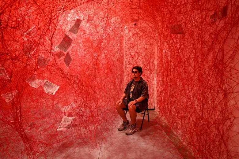 A person sits on a chair in a room filled with dense red string webs and suspended sheets of paper, under red lighting.