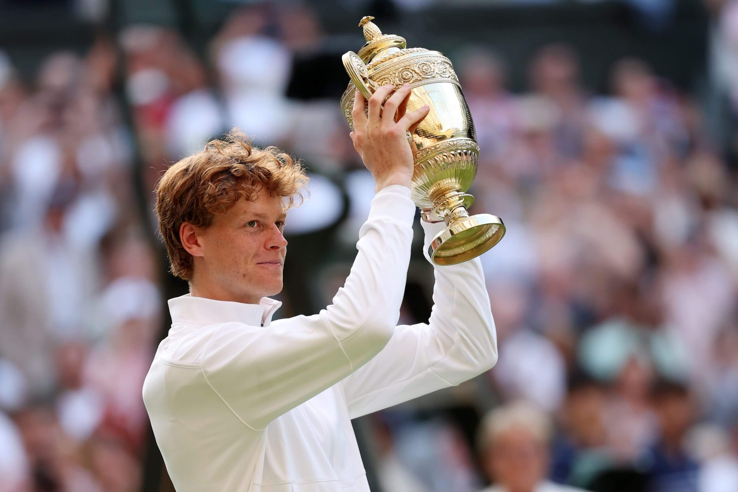 Jannik Sinner holds a golden trophy above his head wearing a white jacket.