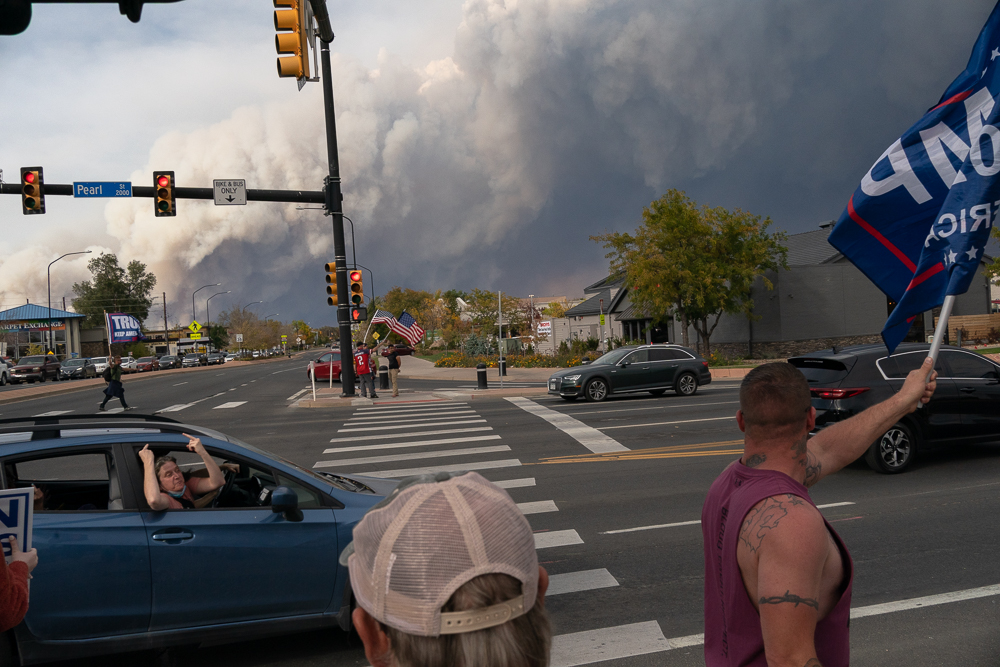 Woman passing in a car raises her middle fingers at flag waving Pro-Trump supporters in Boulder, CO while the CalWood Fire burns in the background on October 17, 2020. On Saturdays leading up to Election Day, November 3, pro-Trump and Republican supporters held gatherings at the southeast corner of 28th and Pearl Streets in Boulder, CO, USA. With a 2019 population of 326,196, Boulder County historically votes Democratic and remains one of the most liberal counties in the state. In the 2004, 2008, 2012, 2016, and 2020 Presidential Elections the Republican Party took only 32%, 26%, 28%, 22%, and 21% of the county vote. The CalWood fire burned 10,106 acres and was the largest fire ever recorded in Boulder County.