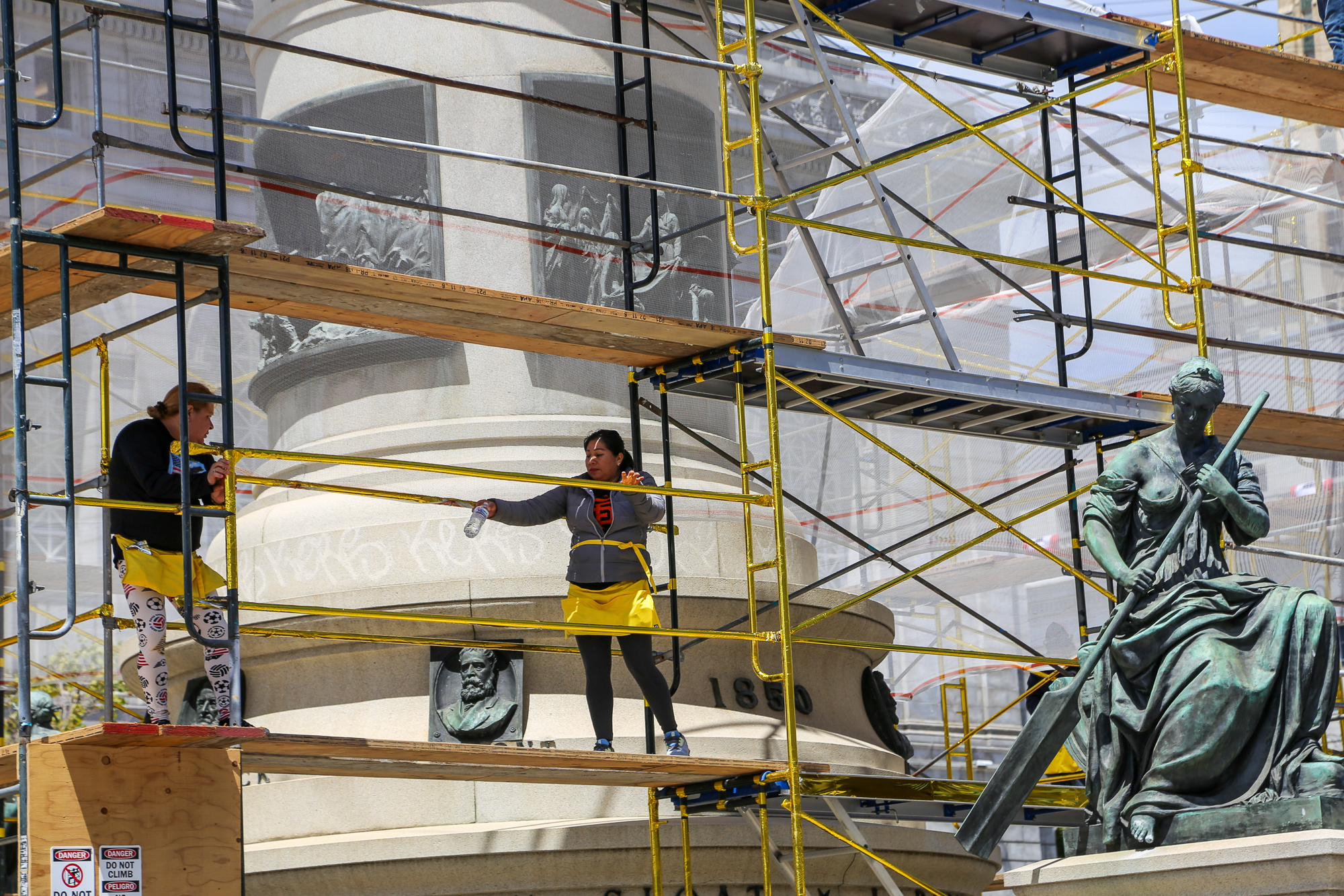 two people stand on scaffolding beside bronze statue