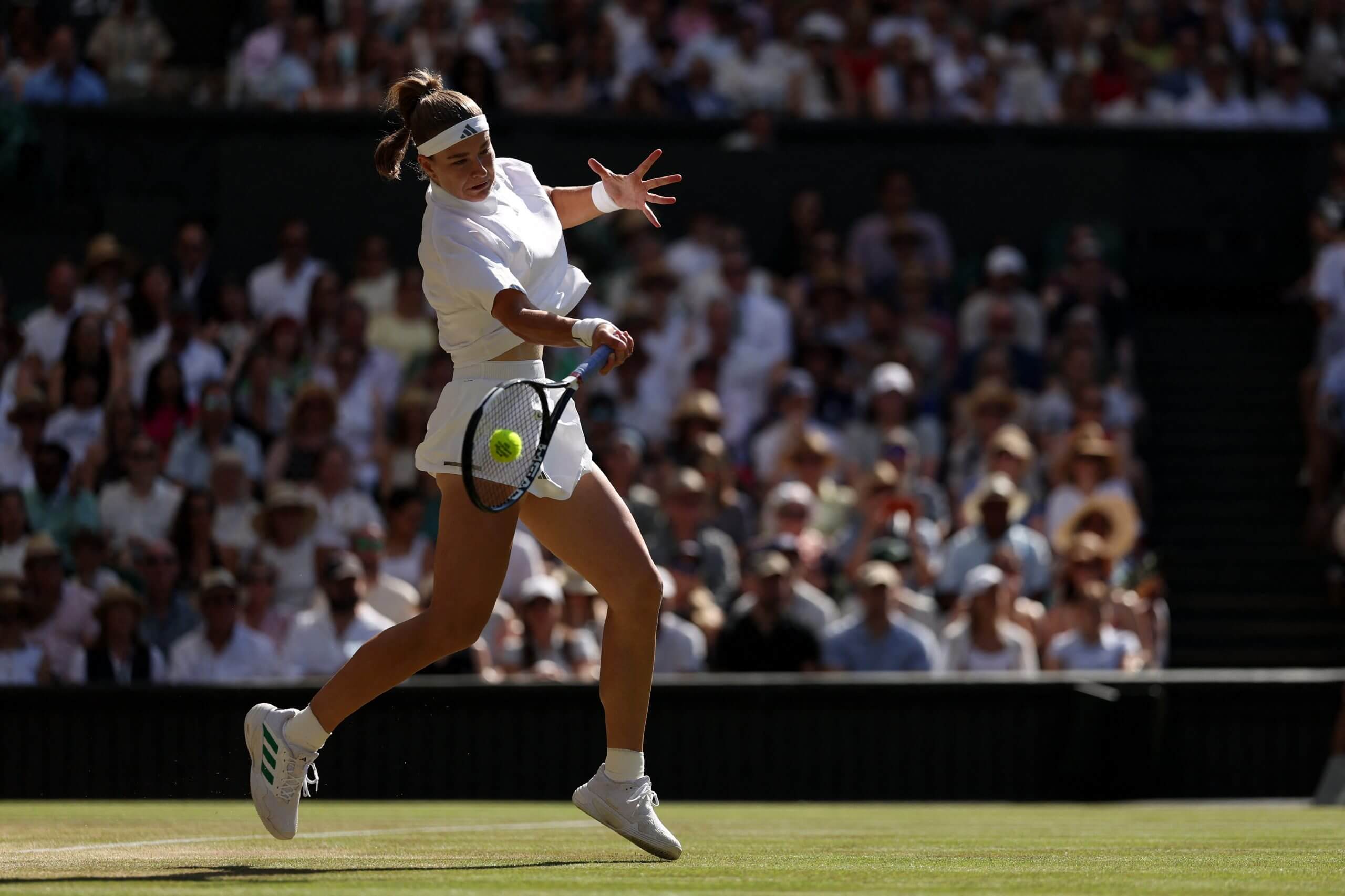 Karolína Muchová hits a forehand on a grass tennis court wearing all-white with a crowd behind her.