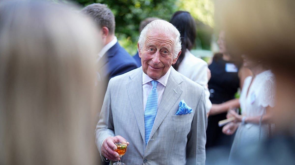 King Charles wearing a beige suit holding a glass as he greets guests at St. James's Palace in London.